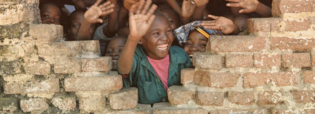 An image of children crowding to an opening in a brick wall to wave as someone goes by, all smiling happily.