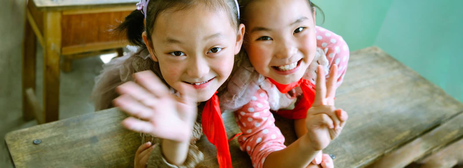An image of two elementary aged girls hugging and smiling up at the camera while in school. One is waving and the other is giving a peace sign.